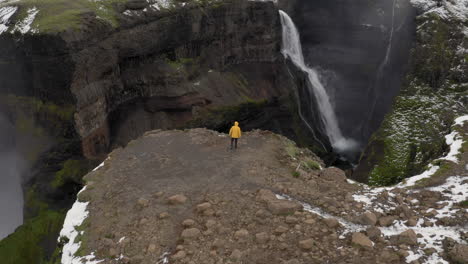 Antenne:-Eine-Person-In-Der-Nähe-Einer-Klippe,-Die-Eine-Gelbe-Jacke-Trägt,-Geht-Auf-Einen-Wasserfall-In-Island-Zu