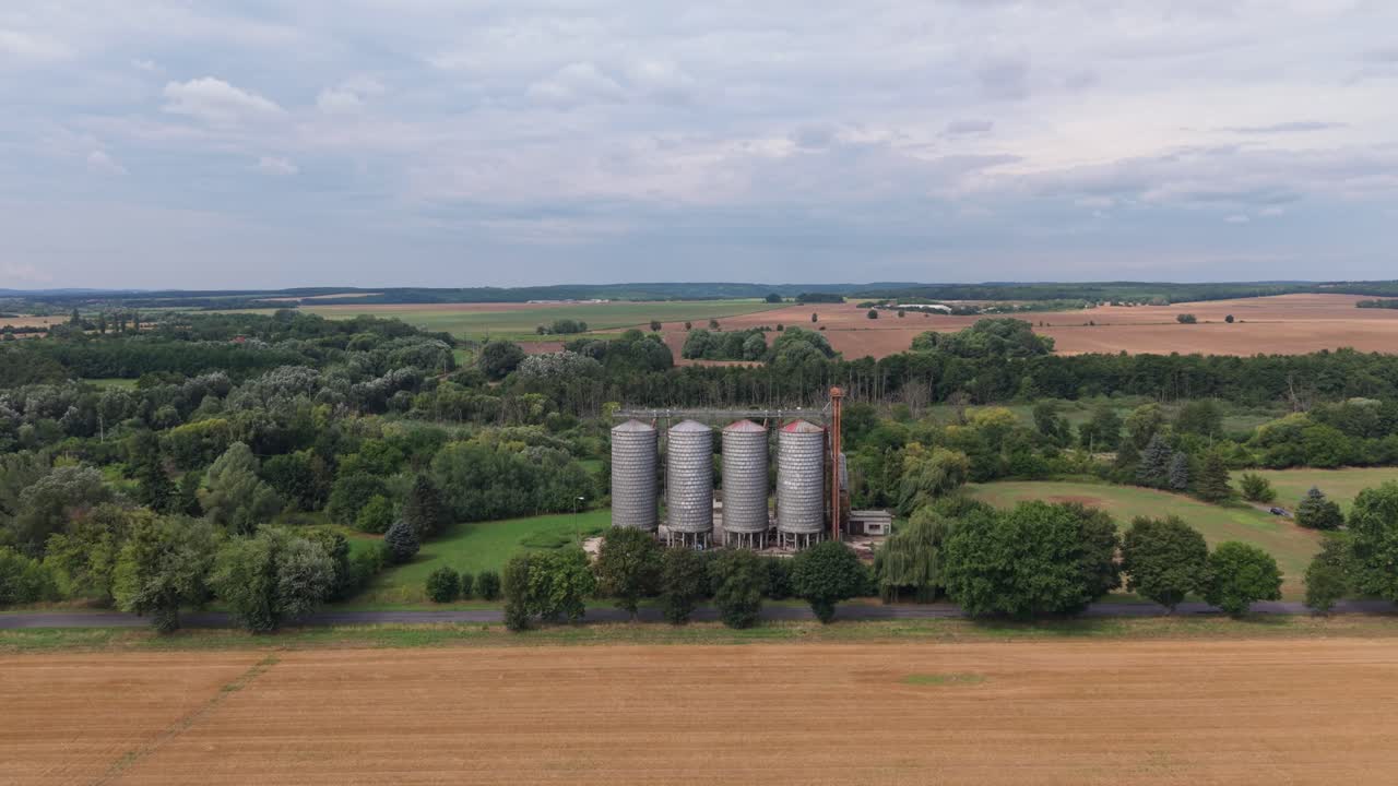 A row of large metal grain silos is situated at the edge of a forested area, surrounded by lush greenery and open farmland. The wide aerial view captures the rural setting under a partly cloudy sky