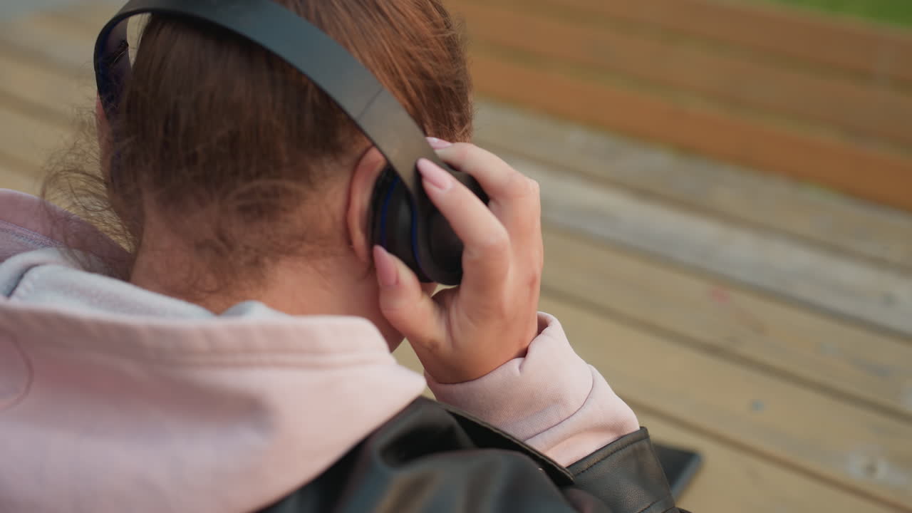 Close up back view of girl in soft pink hoodie and black leather jacket sitting at wooden table slowly putting on headphones as she enjoys cool morning breeze