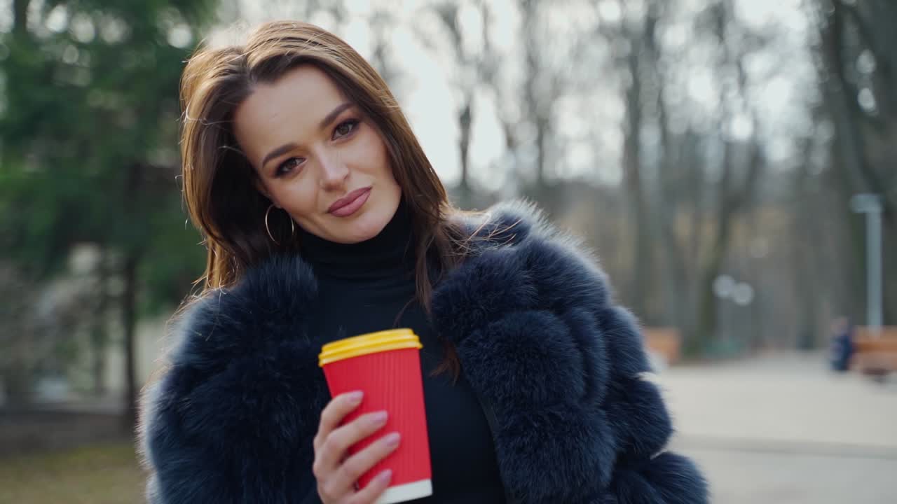 Attractive woman holds coffee in a plastic cup. Portrait of a beautiful girl in fur coat calling someone with her hand in the city park. Slow motion.