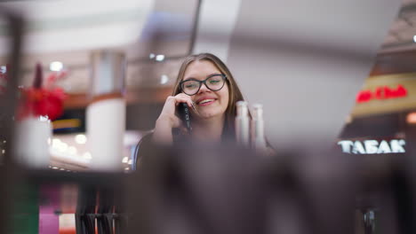 young girl with glasses engaged in phone conversation, looking right, seated at mall food court with blurred background of modern structures, tables, and chairs