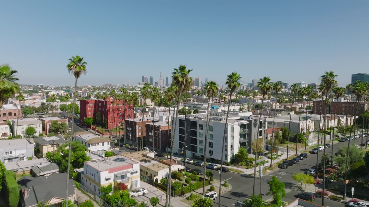 Volo sopra una strada di Los Angeles fiancheggiata da palme in una giornata di sole, ripresa con drone che si affaccia sui tetti con lo skyline di Los Angeles all'orizzonte