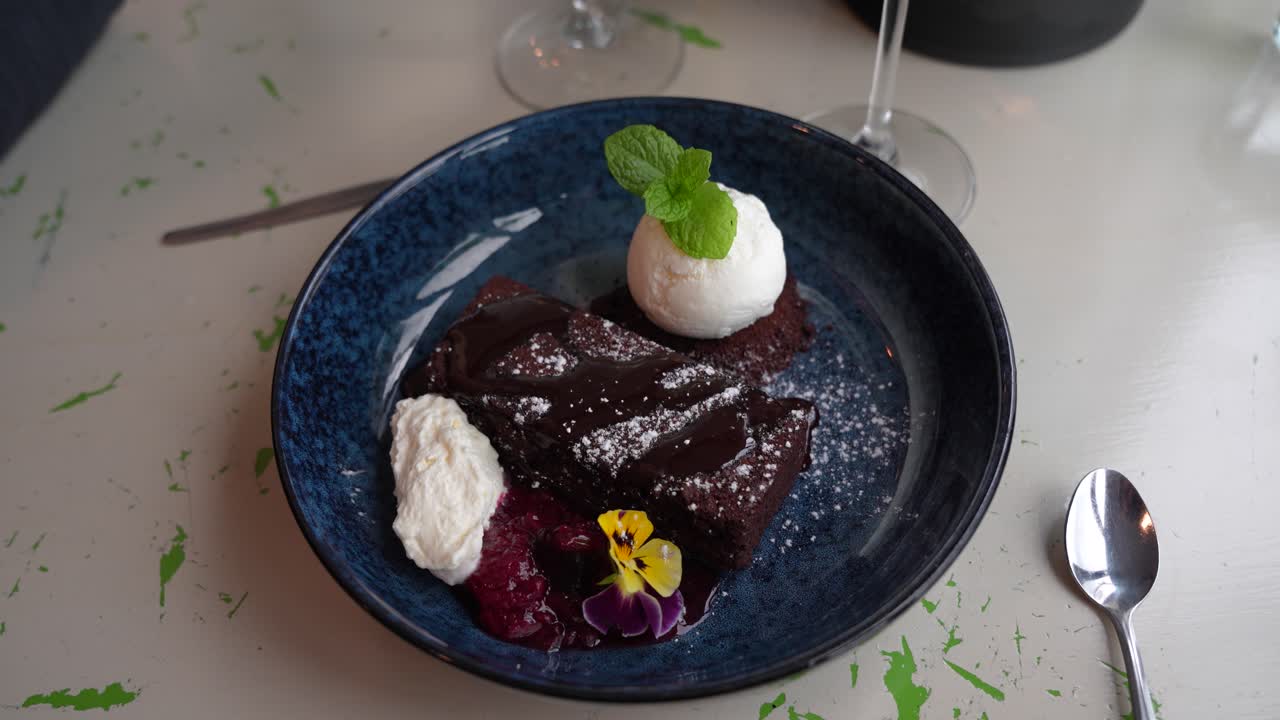 Decadent chocolate brownie with vanilla ice cream, garnished with mint, edible flowers, whipped cream, and berry compote on a blue plate