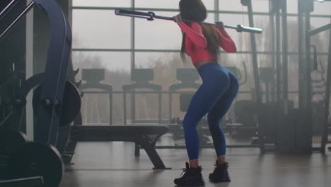 the girl in pink top and blue leggins is doing squats with an empty bar. she is holding the bar behind her neck on shoulders.