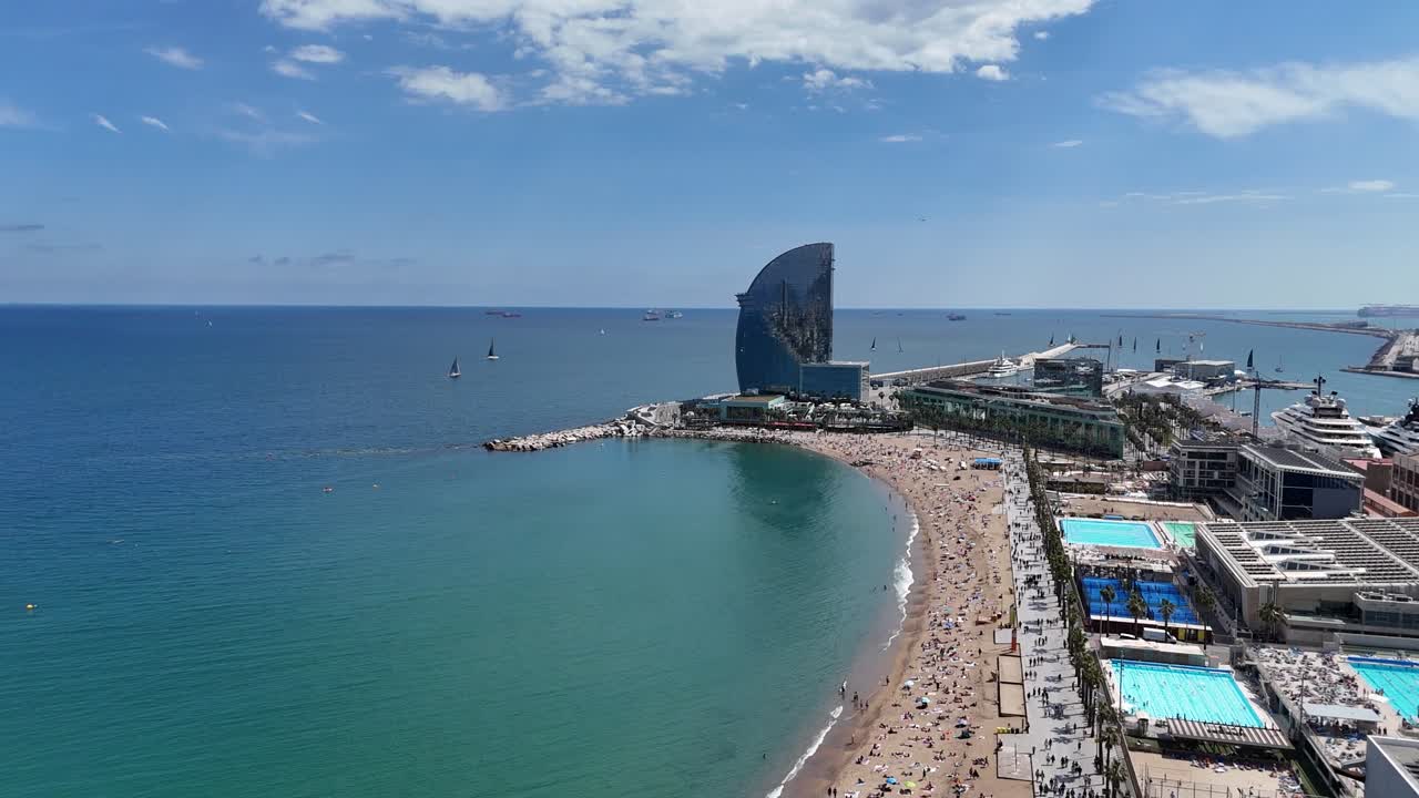 Drone view of Barceloneta Beach with curved sandy shore, clear sea and iconic hotel by water. Swimmers, sunbathers, and modern buildings create vibrant coastal scene in sunny weather