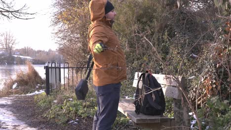 un hombre vestido abrigado quitándose la mochila sentado para descansar en un banco del parque relajándose cerca del lago brooklands, bedford