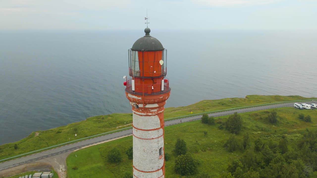 Aerial drone footage close up of a red colored historic lighthouse at a cliff seaside shoreline with a highway underneath it during a misty and foggy day In Pakri Paldiski at summer time. Gorgeous
