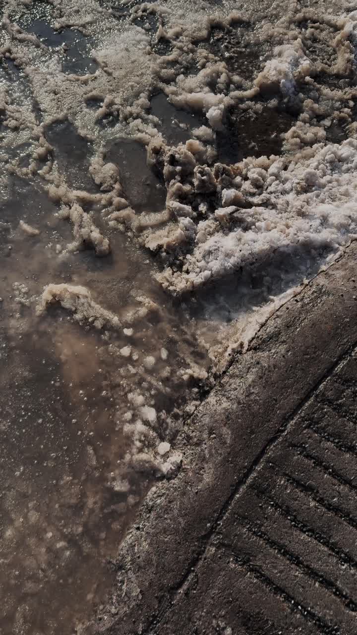 POV coinvolgente di un pedone che attraversa una strada cittadina bagnata con fanghiglia sporca durante il disgelo primaverile. Ottimo per il clima urbano, le transizioni e i social media, formato verticale.