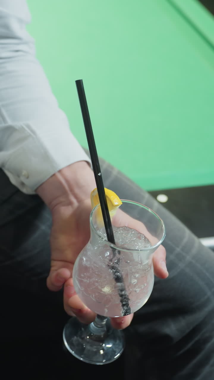 Primo piano di un giovane uomo che indossa una camicia bianca e pantaloni grigi, seduto su un tavolo da biliardo verde, mentre tiene in mano una stecca da biliardo e un bicchiere di limonata con una cannuccia nera e lo solleva per berne un sorso.
