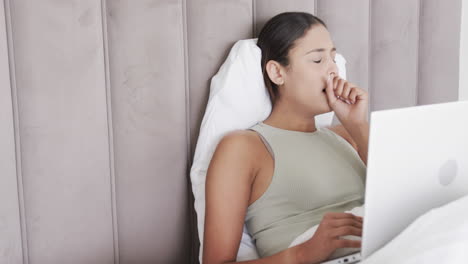 biracial woman sitting on bed using laptop and coughing, slow motion