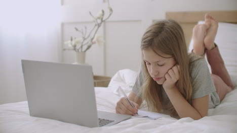 little girl is drawing and writing in copybook lying on bed in front of laptop in her room waiting online lesson