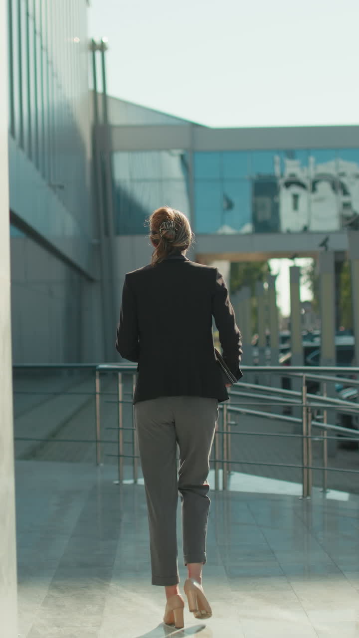 Back view of woman in professional outfit walking past modern residential building with glass facade and parked cars surrounding area in daylight, with shadows cast on clean polished walkway surface
