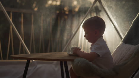 child drinks tea at low table at glamping. tranquil boy rests enjoying beverage sitting on floor in cabin. little kid holds cup near glass wall