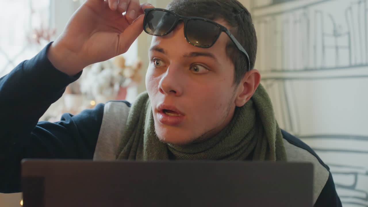 Young caucasian man with laptop peeking over screen, puts on sunglasses and reacts with playful surprise in cozy apartment with bookshelf and fairy lights casual scarf and winter styling add warm