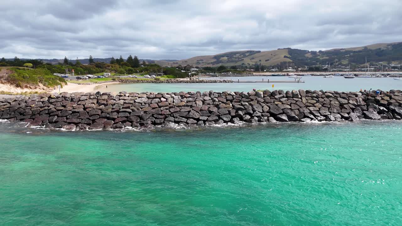 Drone footage captures Apollo Bay's vibrant waters and rocky shoreline under cloudy skies, showcasing the natural beauty of Victoria, Australia