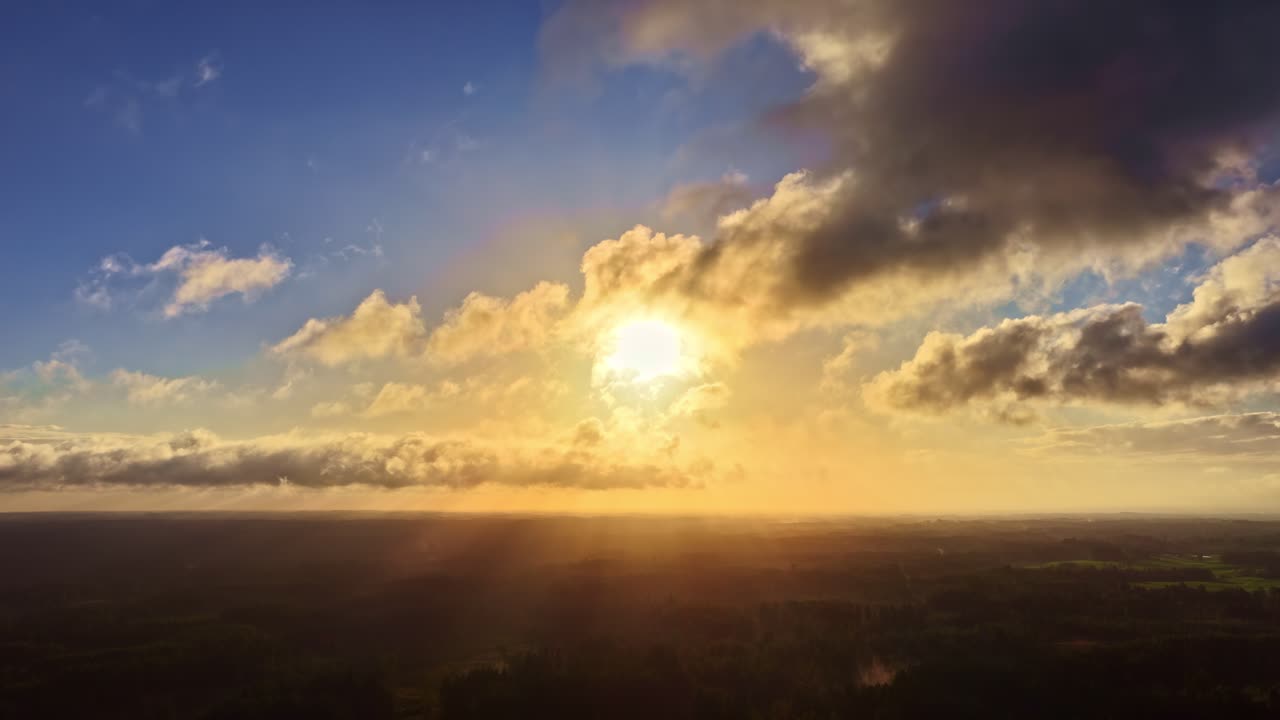 A golden sunrise or sunset lights the sky with clouds passing over a dark, flat landscape