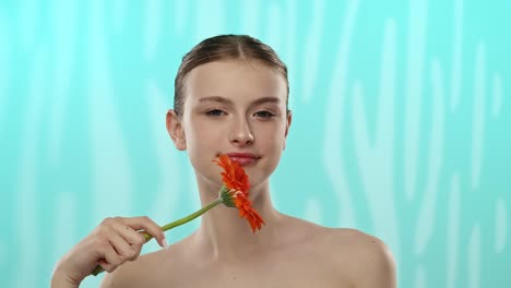 woman posing with orange flower covering eye on blue background a woman holds a vibrant orange flower over her eye against a soft blue patterned backdrop creating a striking visual contrast