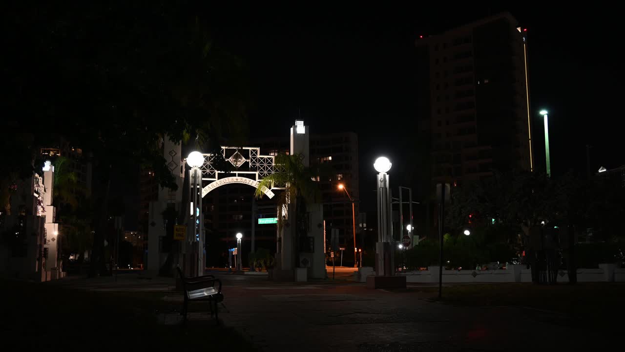 the arch at the entrance of Bayfront park in Sarasota, Florida at night, walkers and traffic go by