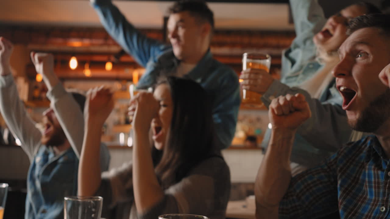 Close-up of a group of fans of men and women sitting together in a bar and watching a broadcast on TV enjoying a goal scored shouting and hugging. in football hockey.