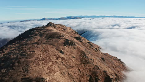 Mountain-peak-rising-above-a-sea-of-clouds-under-clear-blue-skies,-wide-aerial-shot