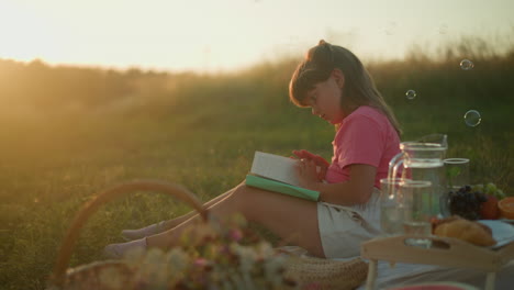 little girl sitting outdoors during golden hour reading book, surrounded by picnic setup with fruits and croissant, warm golden sunlight fills background as bubbles gently float in air