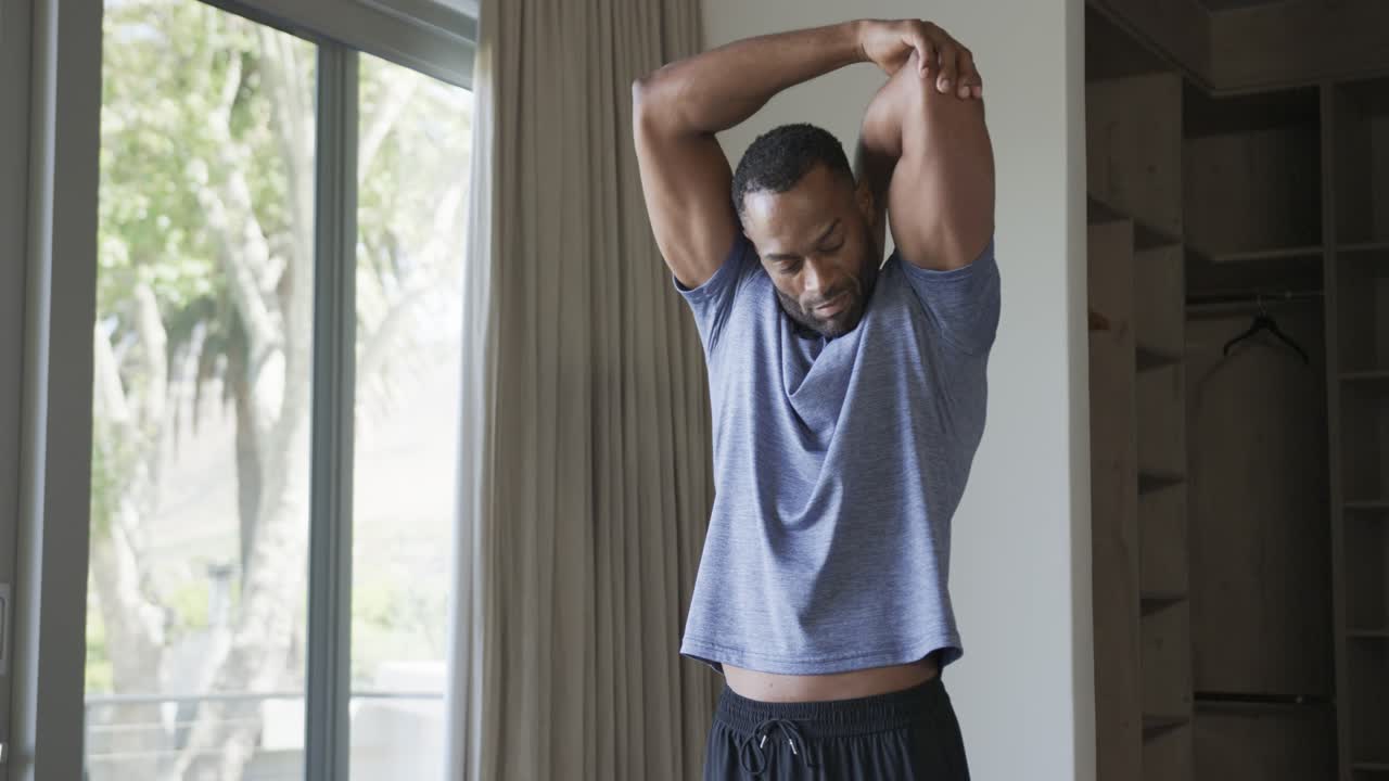 Mature man warming up cross-body to overhead triceps stretch by glass door, blue tee black shorts