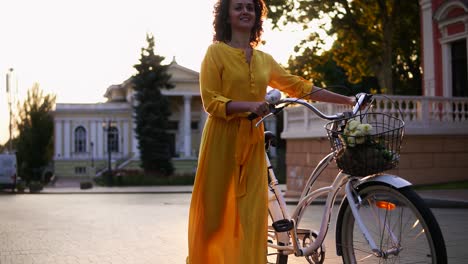 smiling brunette woman in a long yellow dress walking during the dawn holding her city bicycle handlebar with flowers in its