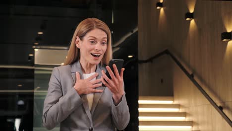 portrait of happy business woman enjoys success on mobile phone in modern office hall near elevator. closeup joyful girl reading good news on phone. surprised female lady celebrating victory on phone.