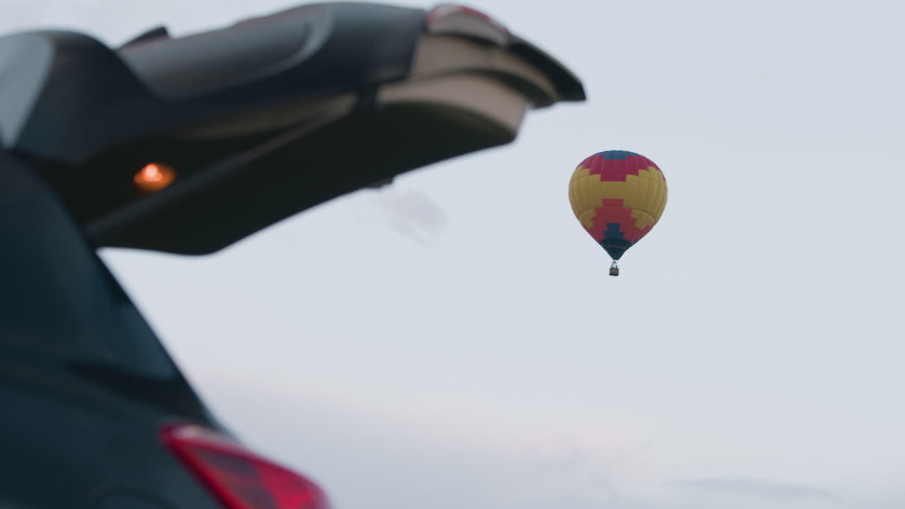 Open car trunk hatch framing distant colorful hot air balloon floating above green grassy field under clear pastel evening sky, trunk bay showing gear and glowing tail light beside parked SUV
