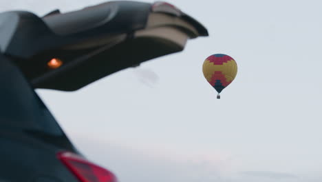 open car trunk hatch framing distant colorful hot air balloon floating above green grassy field under clear pastel evening sky, trunk bay showing gear and glowing tail light beside parked suv