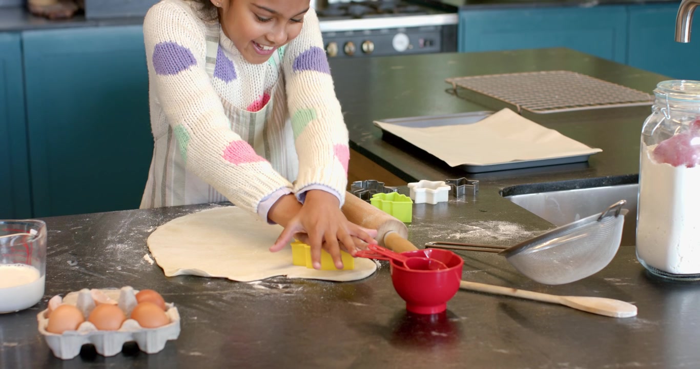 After using rolling pin Indian girl pressing yellow cutter into dough on kitchen island for cookies