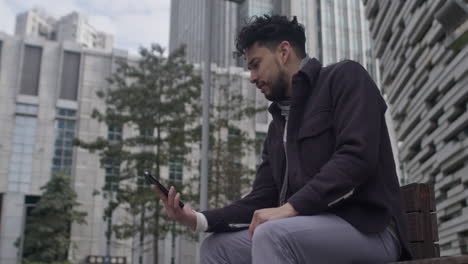 Hispanic-ethnicity-man-dressed-in-warm-casual-clothes-having-a-video-conference-call,-sits-on-the-bench-in-downtown-business-district-in-afternoon