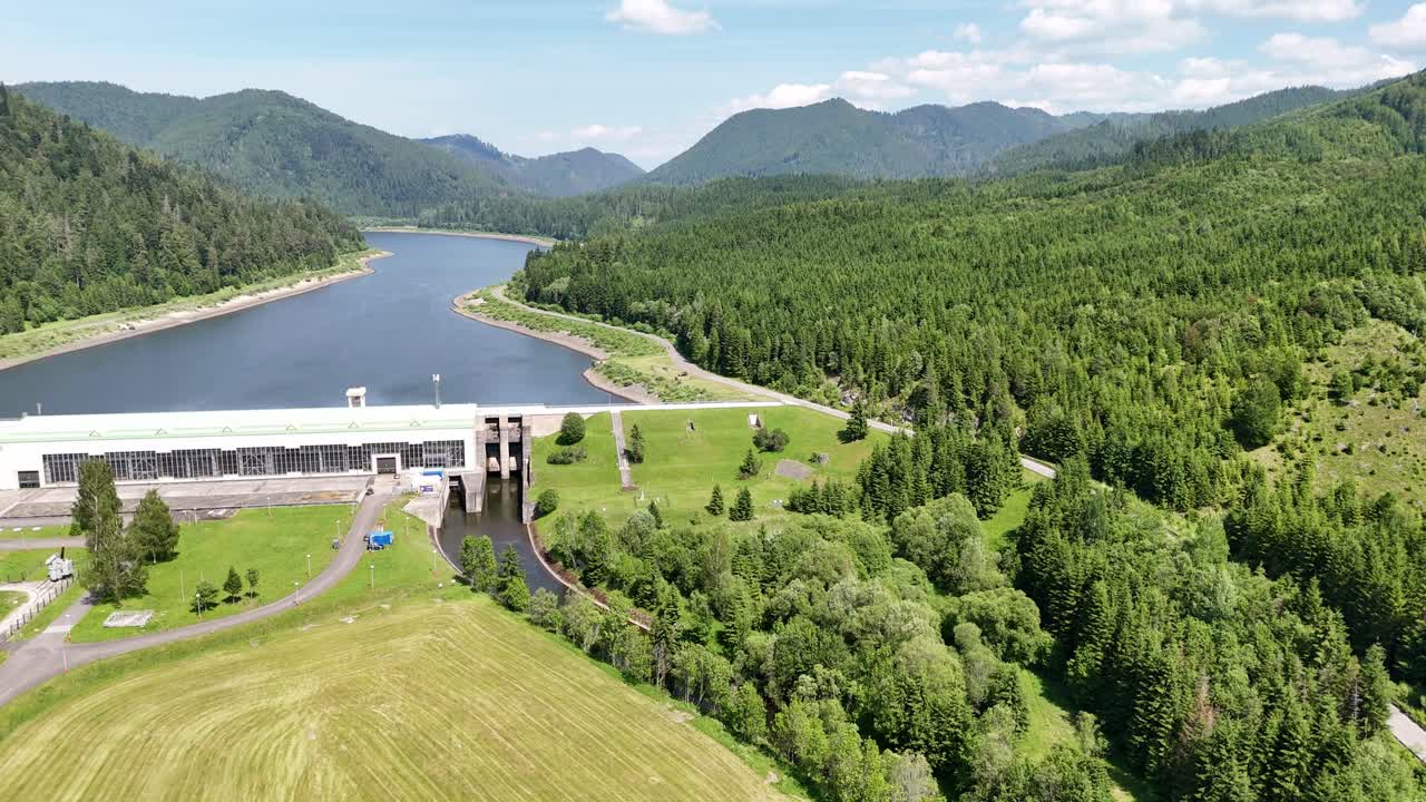 Drone approaching and descending toward Čierny Váh hydroelectric dam, showing the reservoir, power station, and forested mountains under clear summer sky in Slovakia