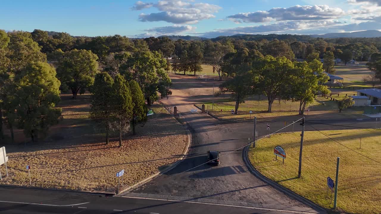 A single vehicle drives along a curved urban street lined with trees and grass, captured in smooth aerial motion during golden hour with long shadows