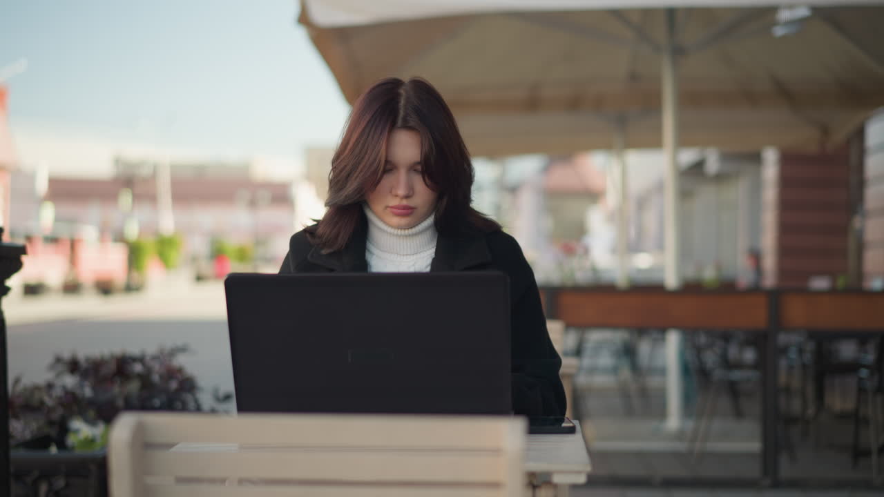 Lady working on laptop outdoors in urban setting, wearing stylish black coat, with decorative black pillar nearby, blurred red brick building in background, and car passing in the blur