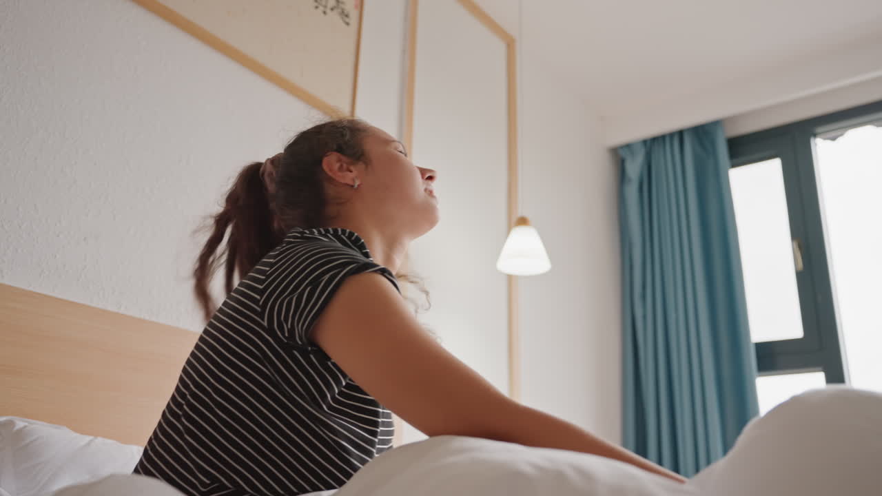 Donna caucasica si sveglia intontita nel letto d'hotel soffocando il rumore con il cuscino, si siede sul bordo del letto socchiudendo gli occhi verso la finestra, piumino in disordine, tende color ottanio, lampada a sospensione, interno moderno e accogliente dell'hotel, frustrazione mattutina