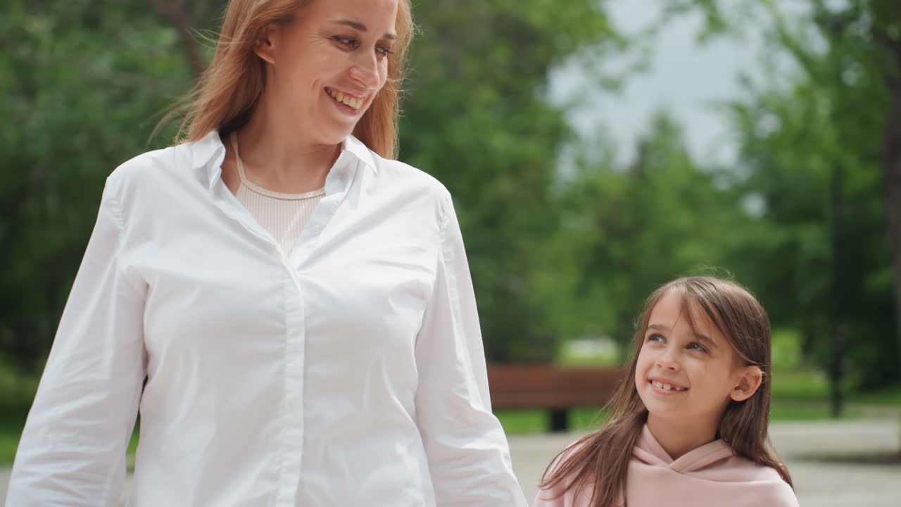 Madre sorride alla figlia durante una passeggiata giocosa nel parco. La bambina si stringe in un abbraccio vicino a panchine e alberi, in un'interazione gioiosa e spontanea. Una connessione calorosa e un'atmosfera rilassata all'aperto.