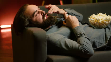 gamer lounged on couch playing videogames on console with bowl of popcorn in lap