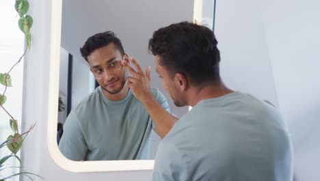 Hombre-Birracial-Feliz-Mirando-En-El-Espejo-Y-Aplicando-Crema-Facial-En-El-Baño,-Cámara-Lenta