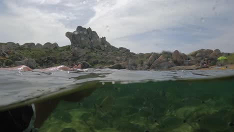 fpv half underwater pov of male legs relaxing while floating on sea water with fish swimming and rocks in background at cala della chiesa cove on lavezzi island in corsica, france