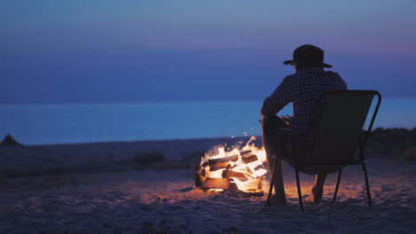 a young man is warming his hands by the fire in the late evening sits by the lake at dusk