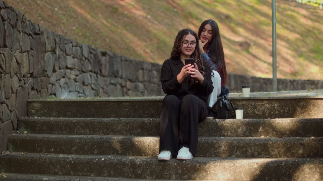 Two young women socializing and using a phone on park stairs