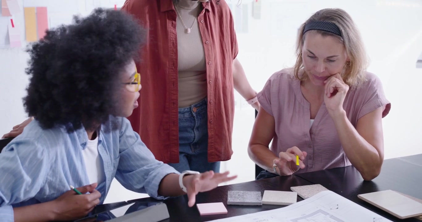 Três mulheres iniciando uma análise de projeto, debruçadas sobre uma planta baixa, selecionando acabamentos com um lápis amarelo