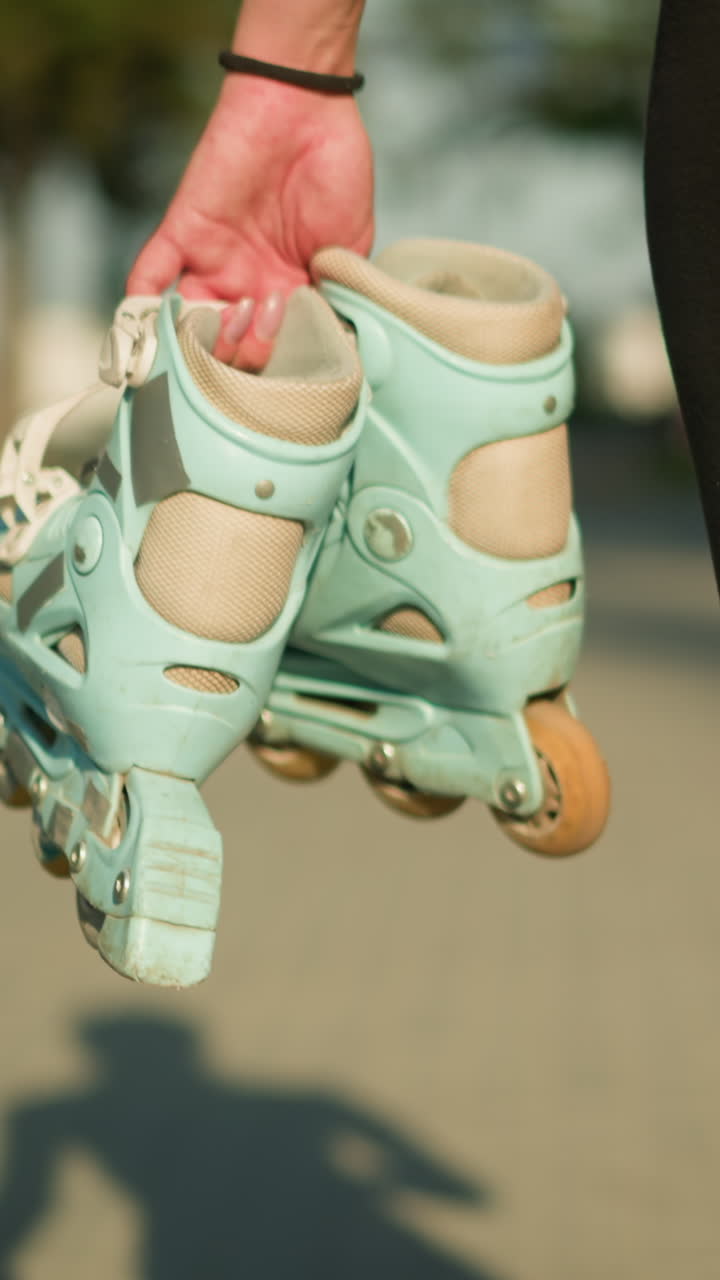 Lower view of lady walking outdoors on interlocked path holding cyan roller skates in her right hand, shadow of her figure is clearly visible on ground, with greenery in the distance background