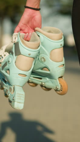 lower view of lady walking outdoors on interlocked path holding cyan roller skates in her right hand, shadow of her figure is clearly visible on ground, with greenery in the distance background