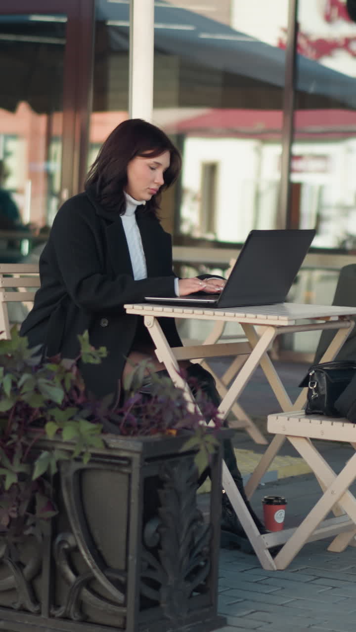 Woman working on laptop outdoors at restaurant, typing at table with plants and coffee cup nearby, focused on task, with blurred background