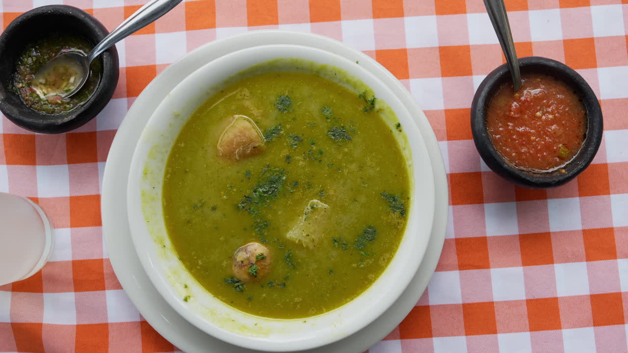 Lähikuva kolumbialaisesta Sancocho de Gallinasta kirkkaalla ruudullisella pöytäliinalla. Täyteläisen kanakeiton lisukkeena mustissa kulhoissa tarjoiltuja annoksia. Eloisat värit ja tekstuurit näkyvillä.