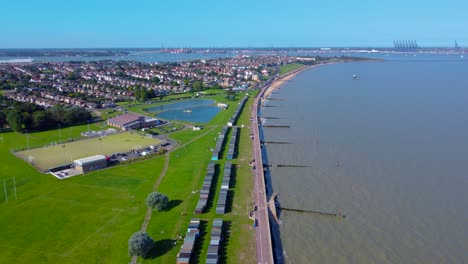 Impresionante-Toma-Costera-Con-Drones-De-Coloridas-Cabañas-De-Playa-De-Dovercourt,-Exuberantes-Campos-Deportivos-Verdes-Y-Un-Sereno-Lago-Con-Botes