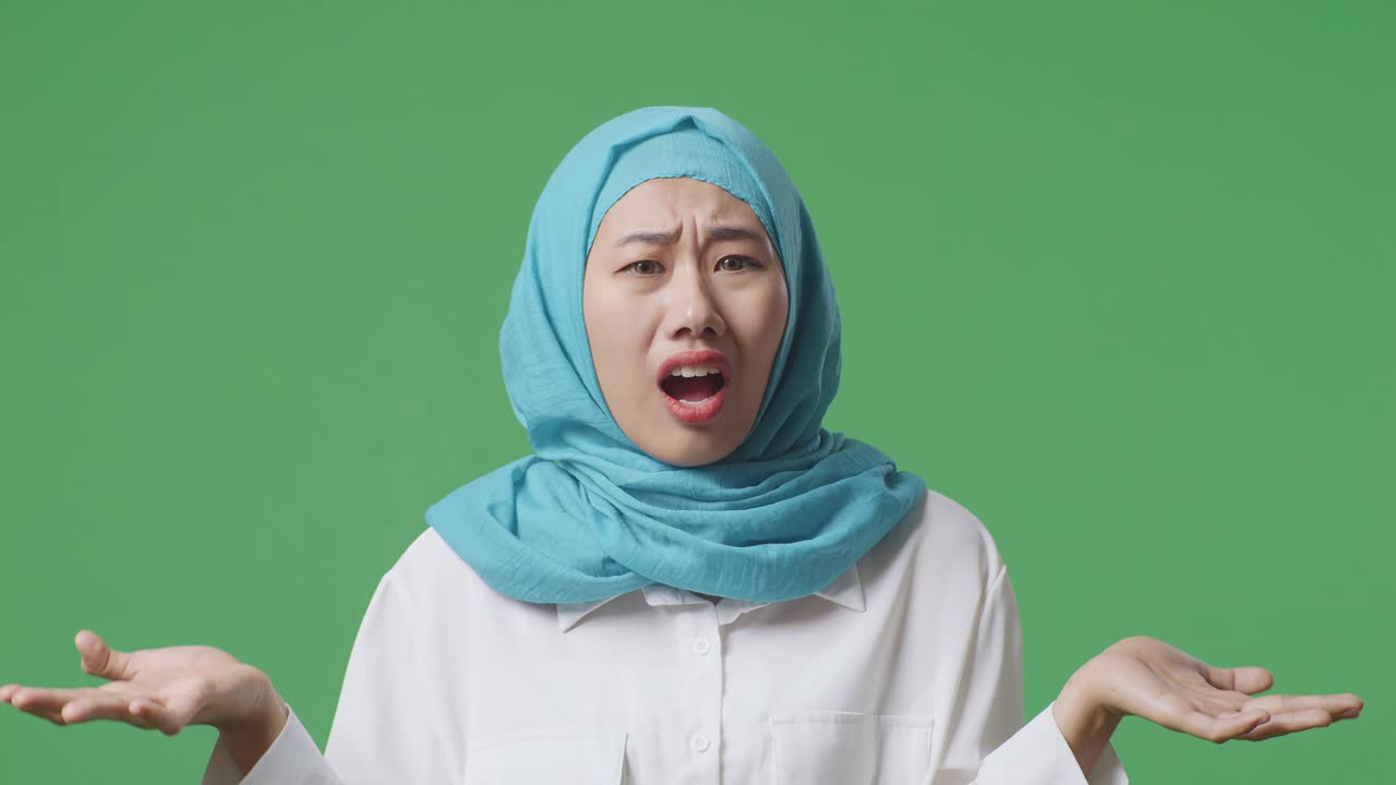 "아시안 무슬림 여성이 ""왜?""라고 묻고 녹색 스크린 배경 스튜디오에서 의심스럽게 서 있습니다."