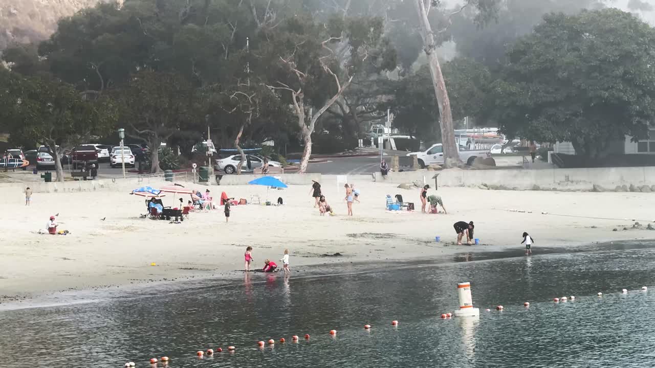 people enjoying Dana Point Beach on a foggy day children playing in the water's white sand with surfers and cabanas in the background hazy surroundings STATIC